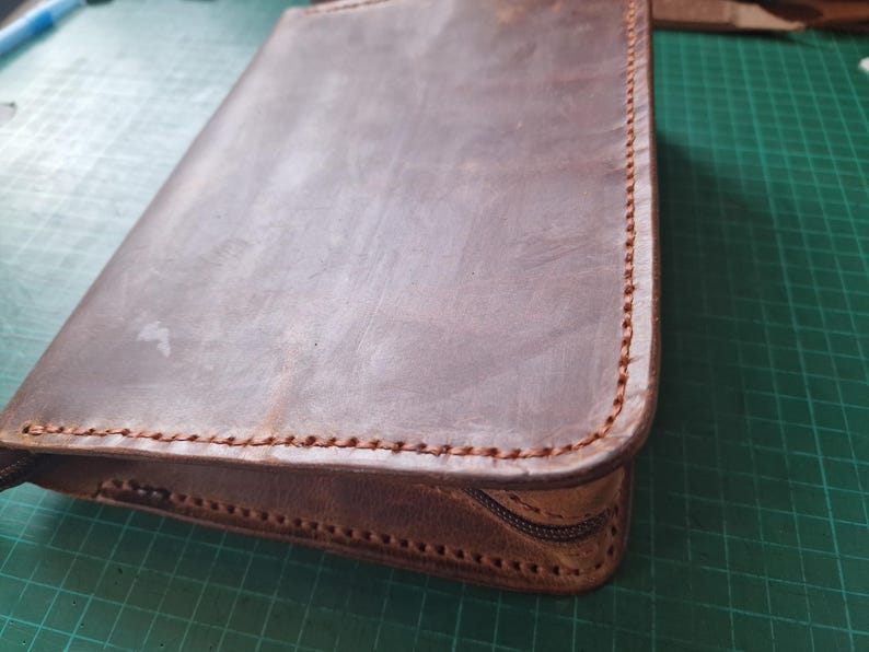 Puede incluir: Un estuche rectangular de cuero marr&oacute;n con bordes cosidos a mano con hilo grueso. El estuche presenta una textura de superficie lisa y mate, junto con una cremallera de color marr&oacute;n oscuro en el lateral para cerrar el contenido de forma segura.