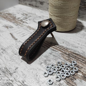 May include: A black leather pull handle with brown stitching and a silver metal button. The handle is attached to a wooden surface with a nail.