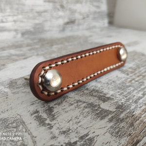 Pode incluir: Uma puxador de gaveta de couro marrom com costuras brancas e duas maçanetas de metal prateado.