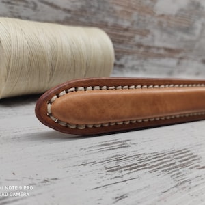 Può includere: Un primo piano di una maniglia in pelle marrone con cuciture bianche. La maniglia è su una superficie di legno.