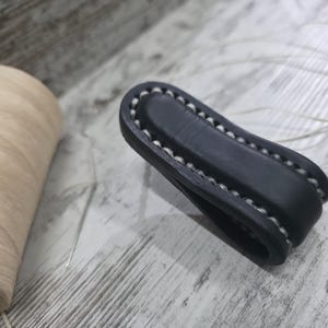 May include: A black leather belt loop keeper featuring thick, light-colored hand-stitched detailing along the edges. The folded leather piece is positioned next to a spool of light beige thread and a thin sewing needle on a light surface.