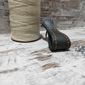 May include: A dark green leather handle with a brown stitch detail. The handle is on a white wooden surface with a spool of beige thread and a pile of silver nuts.
