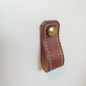 Op de afbeelding: Een bruine leren trekknop met een messingknop. De knop is gestikt met wit garen.
