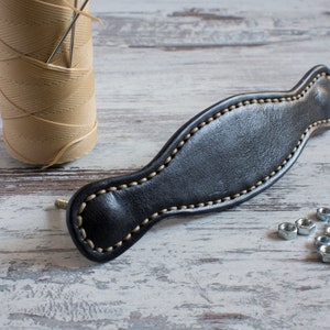 May include: A black leather drawer pull with a decorative stitch detail. The pull is on a white wooden surface with a spool of thread and needles.
