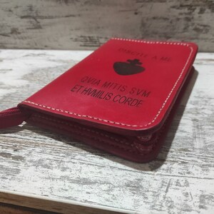 Puede incluir: Una funda de cuero rojo con cremallera, un icono de coraz&oacute;n negro y el texto en lat&iacute;n "DISCITE A ME QVIA MITIS SVM ET HVMILIS CORDE" grabado en la parte frontal. La funda tiene costuras blancas visibles en los bordes y un tirador rojo.