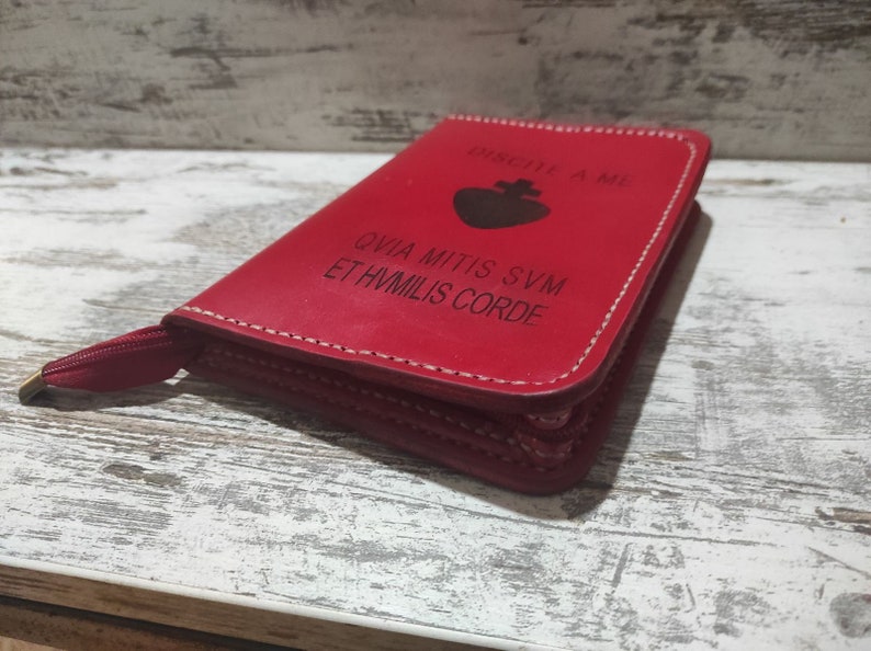 Puede incluir: Una funda de cuero rojo con cremallera, un icono de coraz&oacute;n negro y el texto en lat&iacute;n "DISCITE A ME QVIA MITIS SVM ET HVMILIS CORDE" grabado en la parte frontal. La funda tiene costuras blancas visibles en los bordes y un tirador rojo.