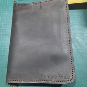 Puede incluir: Una funda de cuero marr&oacute;n oscuro para cuaderno con cierre de cremallera y costuras visibles en los bordes. El nombre Yvonne Walz est&aacute; grabado en la esquina inferior derecha de la portada, con una textura suave y variaciones naturales.