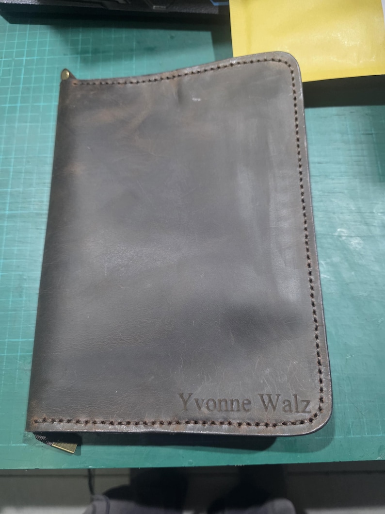 Puede incluir: Una funda de cuero marr&oacute;n oscuro para cuaderno con cierre de cremallera y costuras visibles en los bordes. El nombre Yvonne Walz est&aacute; grabado en la esquina inferior derecha de la portada, con una textura suave y variaciones naturales.