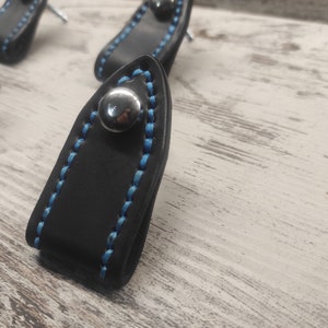 May include: Close-up of a black leather cabinet knob with blue stitching and a silver metal accent. The knob is on a white wooden surface.