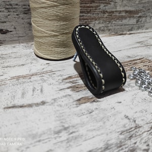 May include: Black leather drawer pull with white stitching. The pull is shaped like a teardrop and has a rounded edge.