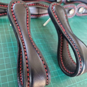 Könnte beinhalten: Zwei schwarze Schubladengriffe aus Leder mit geschlaufenem Design und leuchtend roten Nähten an den Kanten. Jeder Griff verfügt über einen silbernen Metallbefestigungsstift auf der Rückseite, der einen sicheren Halt an Möbeln bietet.