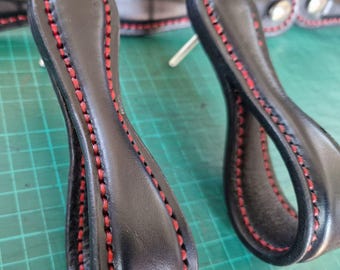 Puede incluir: Dos tiradores de cajón de cuero negro con un diseño en bucle y costuras rojas vibrantes a lo largo de los bordes. Cada tirador cuenta con un poste de montaje de metal plateado en la parte posterior para una fijación segura en muebles.