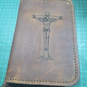 Puede incluir: Una funda de cuero marr&oacute;n con una imagen grabada con l&aacute;ser de un crucifijo con la figura de Jes&uacute;s y las letras INRI en la parte superior. La funda tiene costuras visibles en los bordes y un tirador de cremallera met&aacute;lico.