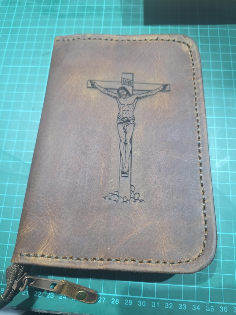Puede incluir: Una funda de cuero marr&oacute;n con una imagen grabada con l&aacute;ser de un crucifijo con la figura de Jes&uacute;s y las letras INRI en la parte superior. La funda tiene costuras visibles en los bordes y un tirador de cremallera met&aacute;lico.