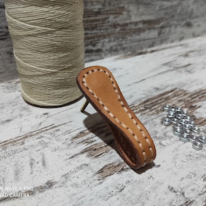May include: A brown leather handle with white stitching. The handle is attached to a wooden surface with a small metal screw.