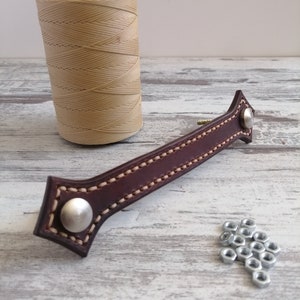 May include: Brown leather handle with a stitched edge and silver rivets. A spool of beige waxed thread and silver nuts are also in the image.