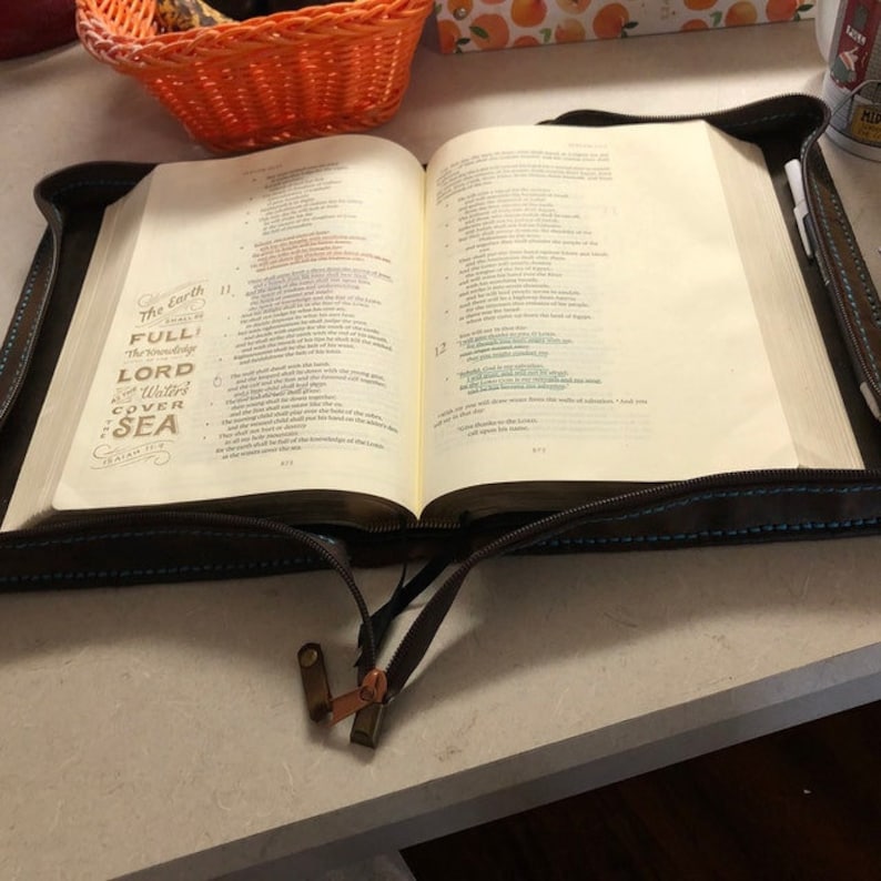 Puede incluir: Una Biblia abierta con una cubierta marr&oacute;n oscura con cremallera y costuras azules. La p&aacute;gina izquierda muestra el texto The Earth shall be full of the knowledge of the Lord as the waters cover the sea, Isaiah 11:9, en letras decorativas.