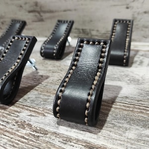 Könnte beinhalten: Fünf schwarze Leder-Schubladengriffe mit einem beigefarbenen Nähten-Detail. Die Griffe sind rechteckig und haben eine leichte Krümmung.