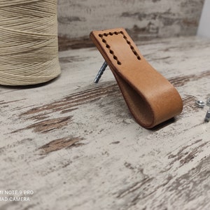 Può includere: Una maniglia a tirare in pelle marrone chiaro con un bordo cucito. La maniglia è su una superficie in legno chiaro con alcune viti sparse intorno.