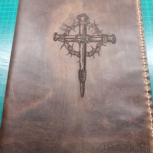 May include: A brown leather journal with a cross and crown of thorns design engraved on the front cover. The name "Donnie Roach" is engraved on the bottom right corner of the cover.