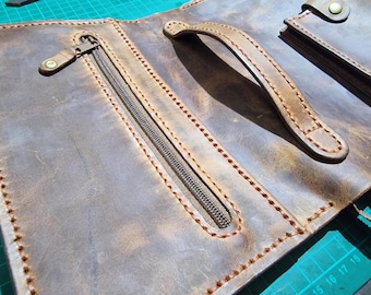 May include: A brown leather portfolio featuring a zippered pocket with a leather pull tab, a sturdy arched handle, and visible thick stitching along the edges. The surface displays a distressed texture with variations in color tones.