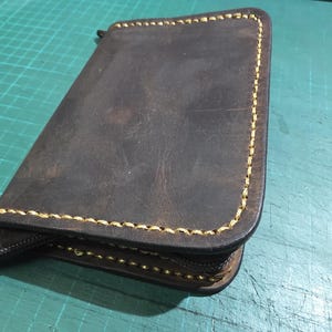 Puede incluir: Una cartera de cuero marr&oacute;n oscuro de forma rectangular con esquinas redondeadas y costuras amarillas gruesas y contrastantes en los bordes. La cartera incluye un cierre de cremallera marr&oacute;n oscuro en el lateral.