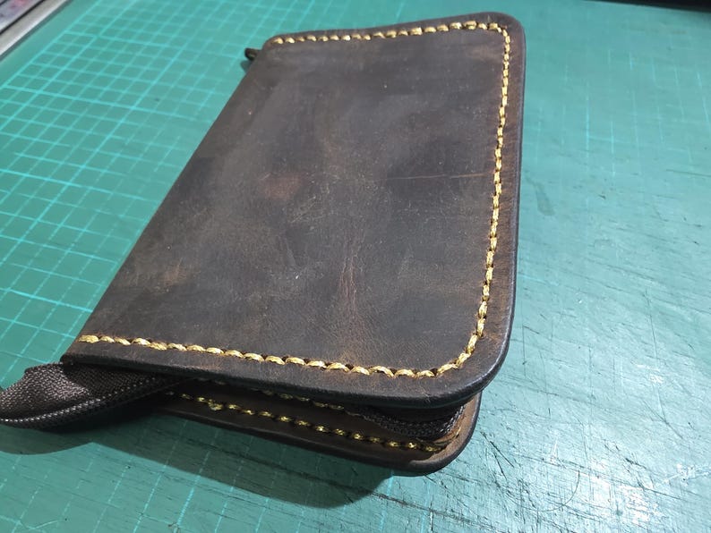 Puede incluir: Una cartera de cuero marr&oacute;n oscuro de forma rectangular con esquinas redondeadas y costuras amarillas gruesas y contrastantes en los bordes. La cartera incluye un cierre de cremallera marr&oacute;n oscuro en el lateral.