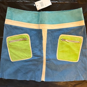 Può includere: Una gonna blu, verde e color crema con due tasche verde lime con cerniere argentate. La gonna ha una fascia in vita turchese e una fascia color crema sotto. Un'etichetta bianca è attaccata alla gonna.