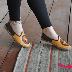 May include: A pair of orange and green leather slippers with a decorative embroidered design. The slippers are on a striped rug.