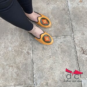 May include: A pair of yellow leather shoes with green and orange embroidered details. The shoes have a rounded toe and a flat sole.