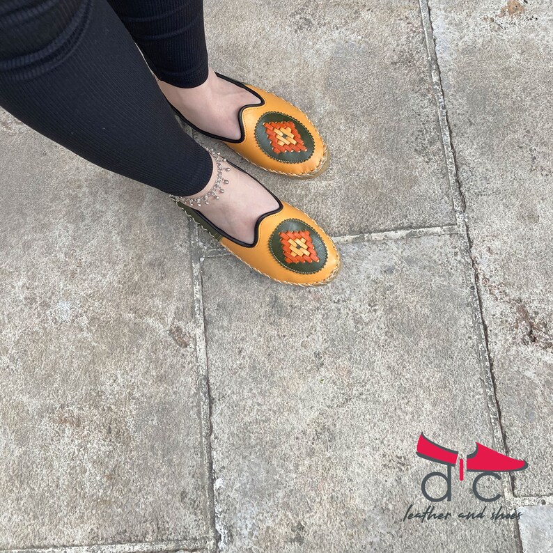 May include: A pair of yellow leather shoes with green and orange embroidered details. The shoes have a rounded toe and a flat sole.