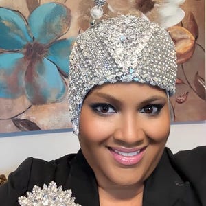 May include: A silver sequined head covering with a matching brooch. The head covering is covered in small, reflective sequins. The brooch is a large, round design with multiple clear crystals. The person is wearing a black blazer.