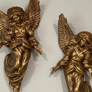 May include: Two gold-colored angel figurines with wings outstretched. The angels are wearing flowing robes and have long hair.