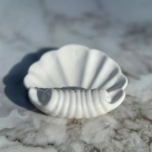 以下が含まれることがあります： 渦巻き模様の装飾が施された白い陶器製の貝殻の形をした皿。ジュエリーや鍵などの小さな物を置くのに最適です。