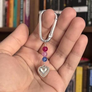 May include: Silver carabiner keychain with a heart-shaped charm and three beads in pink, purple, and blue. The keychain is held in a hand, with a blurred bookshelf in the background.