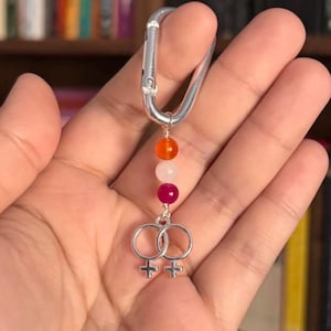 Op de afbeelding: Een zilveren karabijnhaak met een kralen bedel. De bedel heeft oranje, witte en magenta kralen en twee in elkaar grijpende vrouwelijke symbolen. De kralen zijn geregen op een draad. De achtergrond is wazig.