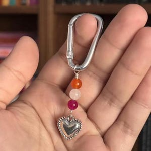 May include: A silver carabiner clip with a heart-shaped charm. The charm has a beaded edge, and a strand of orange, white, and pink beads. The keychain represents the lesbian pride flag.
