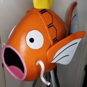 May include: Orange and white fish-shaped sculpture with a yellow crown on top. The sculpture has a large open mouth and black eyes.