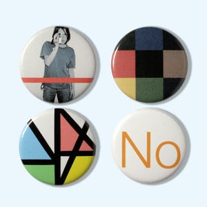 以下が含まれることがあります： 4つの丸いピンバッジ。1つは写真を撮っている人、もう1つはカラフルな市松模様、3つ目は幾何学模様、最後はオレンジ色の「No」という文字が描かれています。