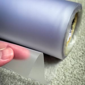 May include: A roll of translucent, frosted plastic film in shades of lavender and grey. A small section of the film is peeled back, revealing its thinness and flexibility. The roll is partially unraveled, showing the inner core.