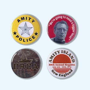 Op de afbeelding: Vier ronde buttons met tekst en afbeeldingen. Eén gele button met "Amity Police" en een ster. Een rode button met een mannengezicht en de quote "We're going to need a bigger boat." De andere buttons zeggen "Orca" en "Amity Island."