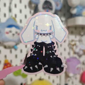 May include: A holographic sticker featuring a white cartoon character with long ears, wearing a spiked collar and large black platform boots with silver spikes. The sticker has a light pink outline and a shimmering, star-patterned finish.