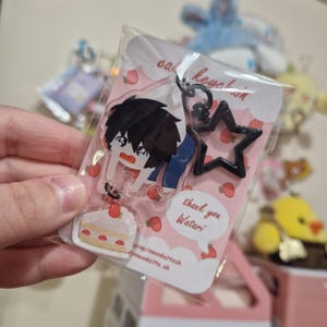 Peut inclure: Un porte-clés en acrylique transparent représentant un personnage de dessin animé avec des cheveux noirs et une chemise bleue, tenant une part de gâteau à la fraise. Le porte-clés est attaché à un charme en forme d'étoile noire. L'arrière-plan est rose avec des fraises blanches et le texte "cake keychain" et "thank you Watari".