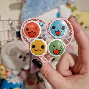 May include: Four colourful round pins with different cartoon faces. The pins are arranged in a heart-shaped holder. The faces are yellow, red, blue, and green.