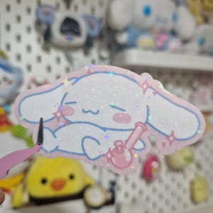 May include: A holographic sticker featuring a white, cartoon-style character with long ears, pink cheeks, and a smiling expression, holding a small pink water pipe. The sticker has a light pink outline and a shimmering, star-patterned finish.