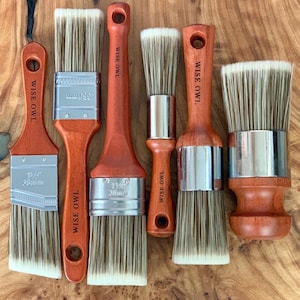 May include: A collection of paint brushes with wooden handles and metal ferrules. The brushes have light-colored bristles and are arranged on a wooden surface. The handles are a reddish-brown color, and the brand name "WISE OWL" is printed on each handle.