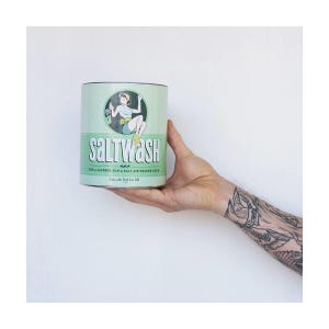 May include: A cylindrical container with the text "SALTWASH" in white, featuring a vintage illustration of a woman in a swimsuit. The container is light green with a dark green rim and base. The text "FOR A LAYERED SUN & SALT AIR SOAKED LOOK" is also visible.