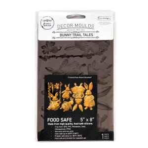May include: A clear plastic package containing a silicone mold with a bunny theme. The mold is dark gray and features various bunny designs, including bunnies on swings and holding carrots. The package is labeled "BUNNY TRAIL TALES" and is food safe, measuring 5" x 8".