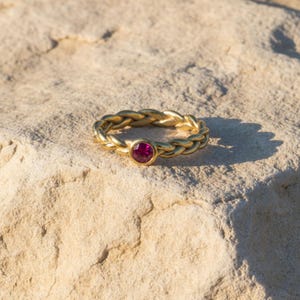 Könnte beinhalten: Ein goldfarbener Ring mit einem geflochtenen Banddesign und einem einzelnen, runden, leuchtend magentafarbenen Edelstein in einer glatten Fassung. Der Ring ist mit einem durchgehenden, gedrehten Metallmuster gestaltet, das den Finger umschließt und den Kontrast zwischen dem goldenen Metallfinish und dem dunkelrosa Stein hervorhebt.