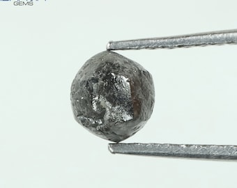 Puede incluir: Un diamante redondo de corte áspero de color gris oscuro sostenido por unas pinzas. El diamante tiene aproximadamente 5 mm de diámetro.